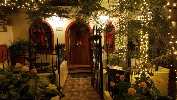 Una entrada cálidamente iluminada, con un sendero de baldosas que conduce a una puerta de madera, flanqueada por plantas en macetas y luces decorativas.