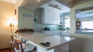 A modern kitchen with white cabinets, a central island with bar stools, a clock on the wall, and a window with blinds.