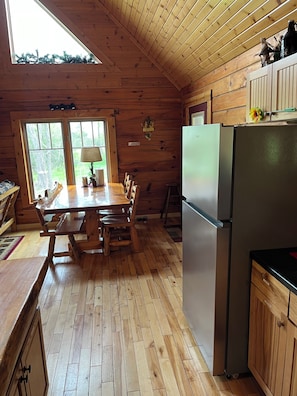 A wooden cabin interior with a dining table, chairs, a refrigerator, and a window with a view of trees. - Lovely Devils Lake Cabin Baraboo Dells Huge Yard Skiing Best Location! (Baraboo)