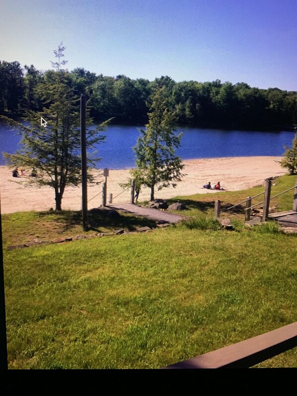 Blick auf einen See mit einer Holzterrasse, einem Sandstrand und einem bewaldeten Ufer im Hintergrund.