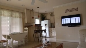 A living room with a television, a dining area with chairs, a kitchen with cabinets and a refrigerator, and a sign that reads "MARCO ISLAND".