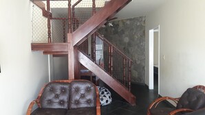 Un escalier avec une rampe en bois et une barrière en treillis métallique. On aperçoit deux fauteuils en cuir et un coussin à motifs au premier plan.