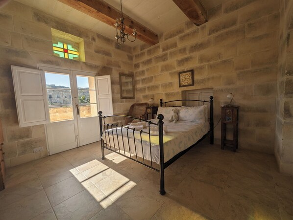 A bedroom with a stone wall, a bed with a teddy bear, a nightstand, and a window with a view. - Large modern farmhouse with private pool & BBQ, stunning views, perfect location (Ir-Rabat Għawdex)