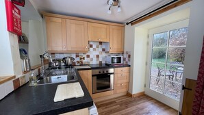 A modern kitchen with wooden cabinets, a black countertop, a stainless steel sink, a microwave, an oven, and a window with a view of the outdoors. - Holly Cottage is ideal for families of 4, with outdoor seating (Taynuilt)