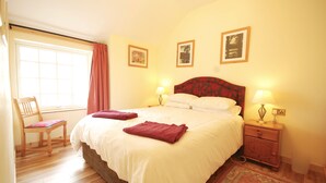 A neatly made bed with two red towels, a wooden chair, a nightstand with a lamp, and three framed pictures on the wall. - Holly Cottage is ideal for families of 4, with outdoor seating (Taynuilt)