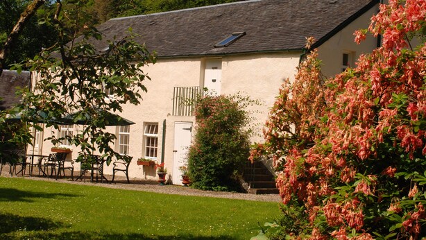 A traditional building with a sloped roof, a small window, and a door. There is a garden with a lawn, a bench, and a potted plant. - Holly Cottage is ideal for families of 4, with outdoor seating (Taynuilt)
