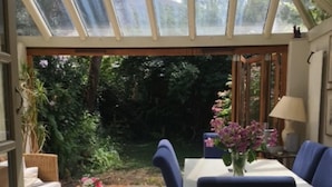 A sunlit patio with a table and chairs, a sofa, and a glass ceiling overlooking a garden.