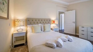 A neatly arranged bedroom with a large bed, two bedside tables, a nightstand, a wardrobe, and a door leading to another room.