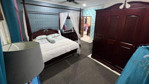 Quarto com cama de dossel, ventoinha no teto, guarda-roupa e banheiro visível por uma porta aberta.