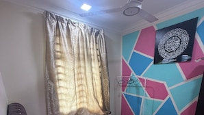Une chambre à coucher avec un lit, des rideaux, un ventilateur de plafond et un téléviseur fixé au mur.
