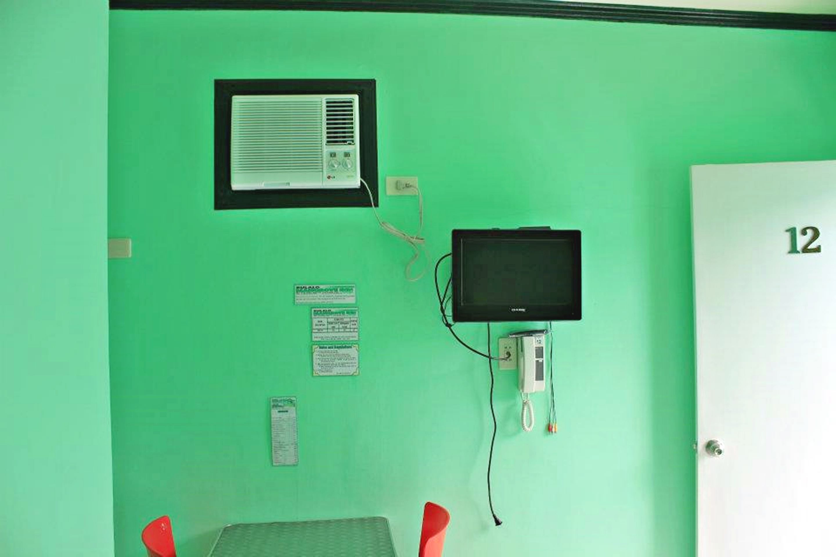 A room with a green wall, a white door with the number 12, a small table with a chair, a television, and a wall-mounted air conditioning unit.
