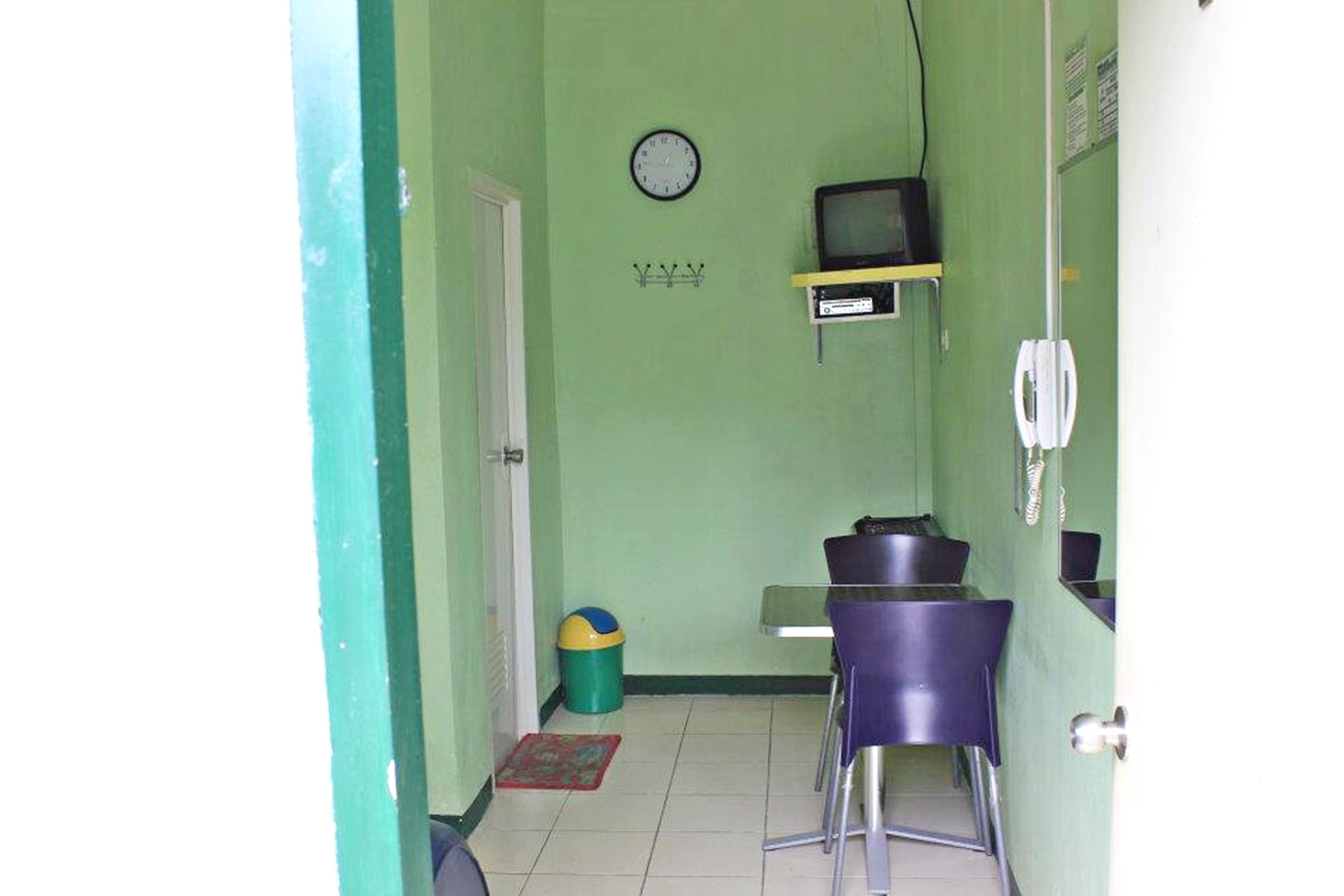 Een kleine kamer met een groene muur, een klok, een televisie op een plank, een prullenbak en een deur.