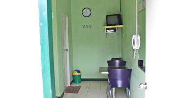 Een kleine kamer met een groene muur, een klok, een televisie op een plank, een prullenbak en een deur.