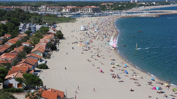 Une plage côtière bordée de bâtiments aux toits de tuiles rouges, une plage de sable fin et une mer d’un bleu limpide.
