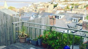 Una terraza de madera con un banco, plantas en macetas y vistas a un pueblo costero.