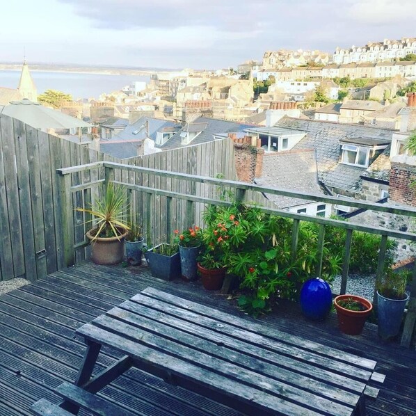 Una terraza de madera con un banco, plantas en macetas y vistas a un pueblo costero.