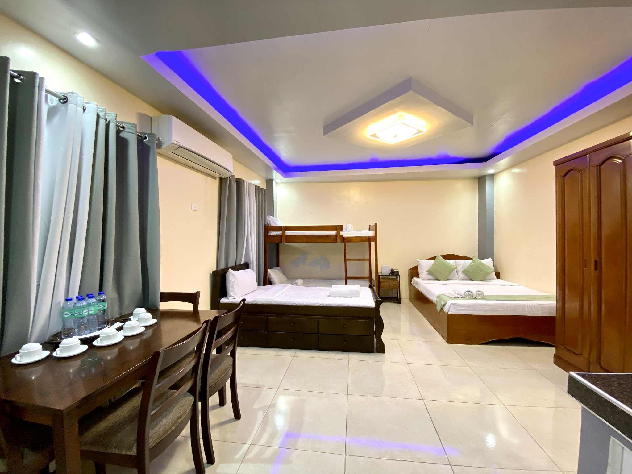 Habitación de hotel con una mesa de comedor para dos personas, una cama con cabecera de madera, una mesita de noche con lámpara y un armario.