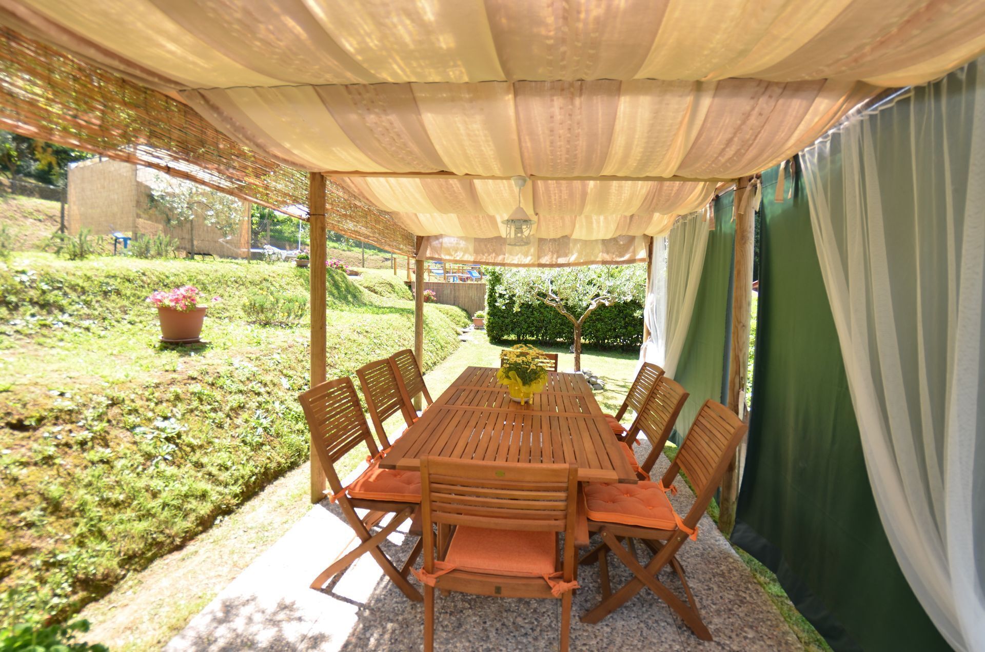 Een terras buiten met een houten tafel en stoelen, een pergola erboven en uitzicht op een tuin.