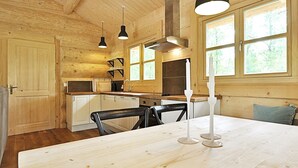 Een houten blokhut van binnen met een eettafel, stoelen, een keuken en grote ramen.