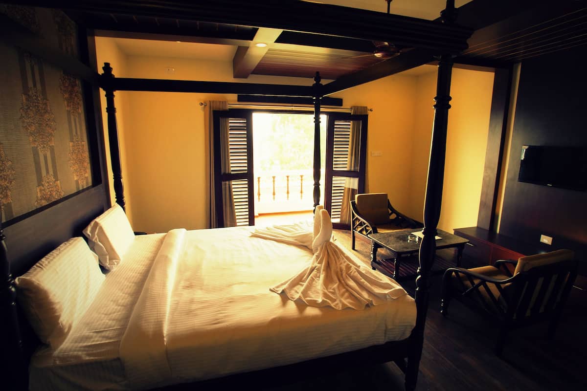 Una cama con dosel con una colcha blanca, cabecera de madera y una silla junto a la ventana.