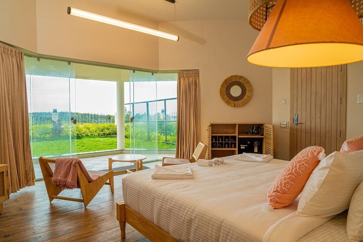 Habitación de hotel con una cama grande, una silla de madera, una mesita pequeña, una estantería y vistas a un paisaje verde a través de amplias ventanas.