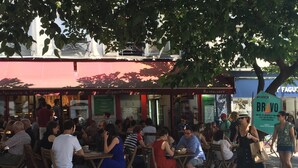 Un café al aire libre con mesas y sillas de madera, rodeado de árboles y un toldo rojo.