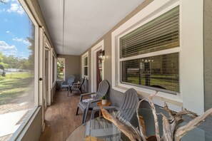 En overdækket veranda med udendørs møbler, store vinduer og udsigt til naturen.