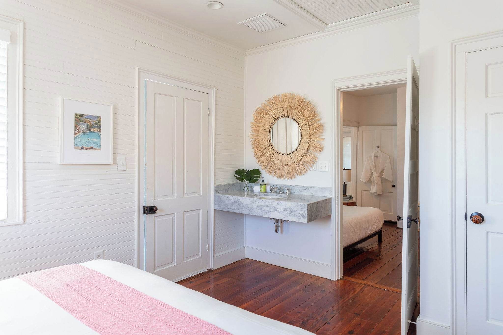A bedroom with a bed, a vanity with a mirror, a picture on the wall, and a door leading to another room.