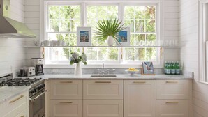 A kitchen with white cabinets, a stove, and a window with a view of greenery. - Casa Zoë | Cabana House – Bold Design, Prime Location (Charleston)