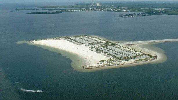 An aerial view of a coastal resort with a long sandy beach, a marina, and a small island with buildings and greenery. - Quiet Gulf Canal Neighborhood with Enclosed Lanai & Heated Pool EV friendly (Hudson)