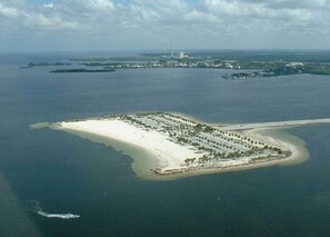An aerial view of a coastal resort with a long sandy beach, a marina, and a small island with buildings and greenery. - Quiet Gulf Canal Neighborhood with Enclosed Lanai & Heated Pool EV friendly (Hudson)