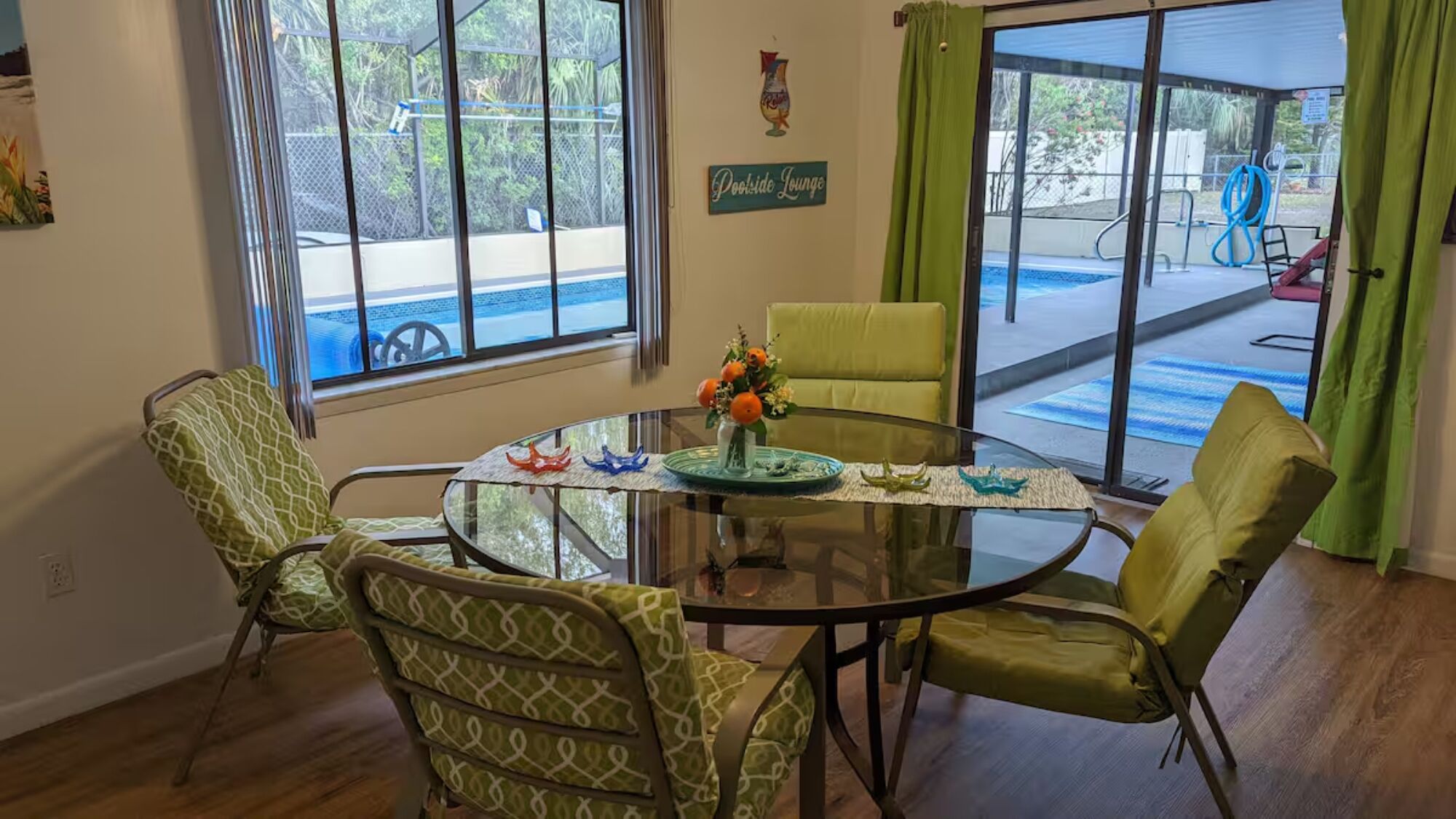 Uma sala de jantar com mesa de vidro, cadeiras estampadas em verde e vista para um espaço de lounge junto à piscina.