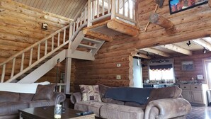 A log cabin interior with a staircase, wooden walls, a living room with a sofa and cushions, and a kitchen area. - Luxury Log Home at Foot of Mt. Princeton (Buena Vista)