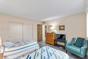 Un dormitorio con dos camas, un aparador de madera, un televisor y un sillón color aguamarina.