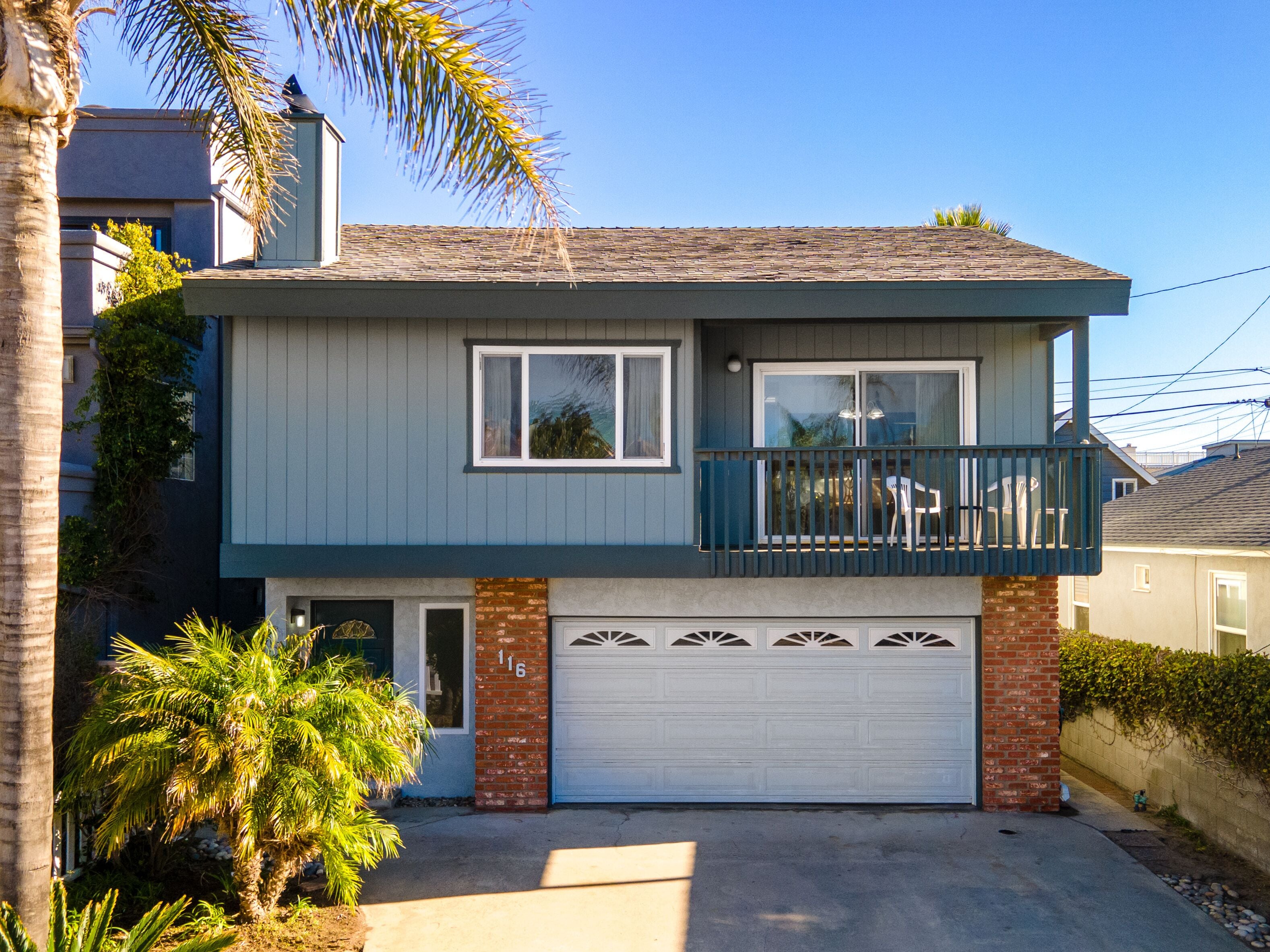 A two-story house with a garage, a balcony, and a small front yard with a palm tree.