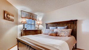 Un dormitorio con una cama de madera, dos mesitas de noche con lámparas, una ventana con persianas y un adorno en la pared.