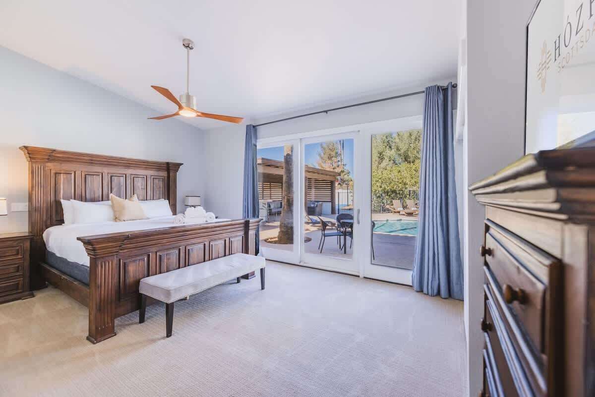 Un dormitorio con cama de madera, un banco, ventilador de techo y vista a una piscina exterior a través de amplios ventanales correderas.