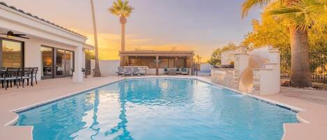 A swimming pool with lounge chairs, a covered seating area, and palm trees.