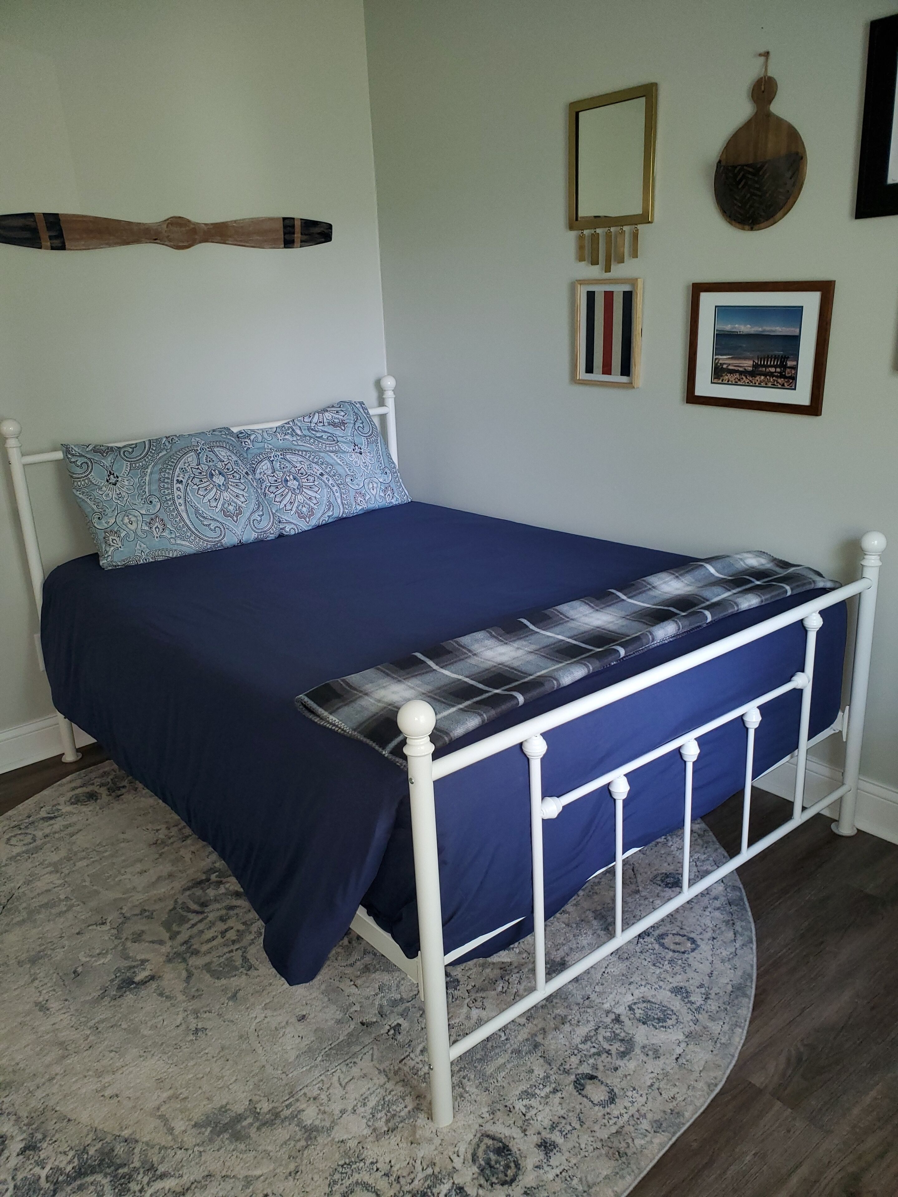 A bedroom with a bed, a patterned pillow, a blue bedspread, a plaid blanket, a round rug, and wall decorations including a mirror, a wooden object, and framed pictures.