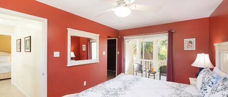 Un dormitorio con cama, ventilador de techo, ventana con cortinas y puerta que da a otra habitación.