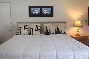 A neatly made bed with a white duvet and patterned pillows, a framed artwork above the bed, and a bedside lamp on a wooden nightstand.