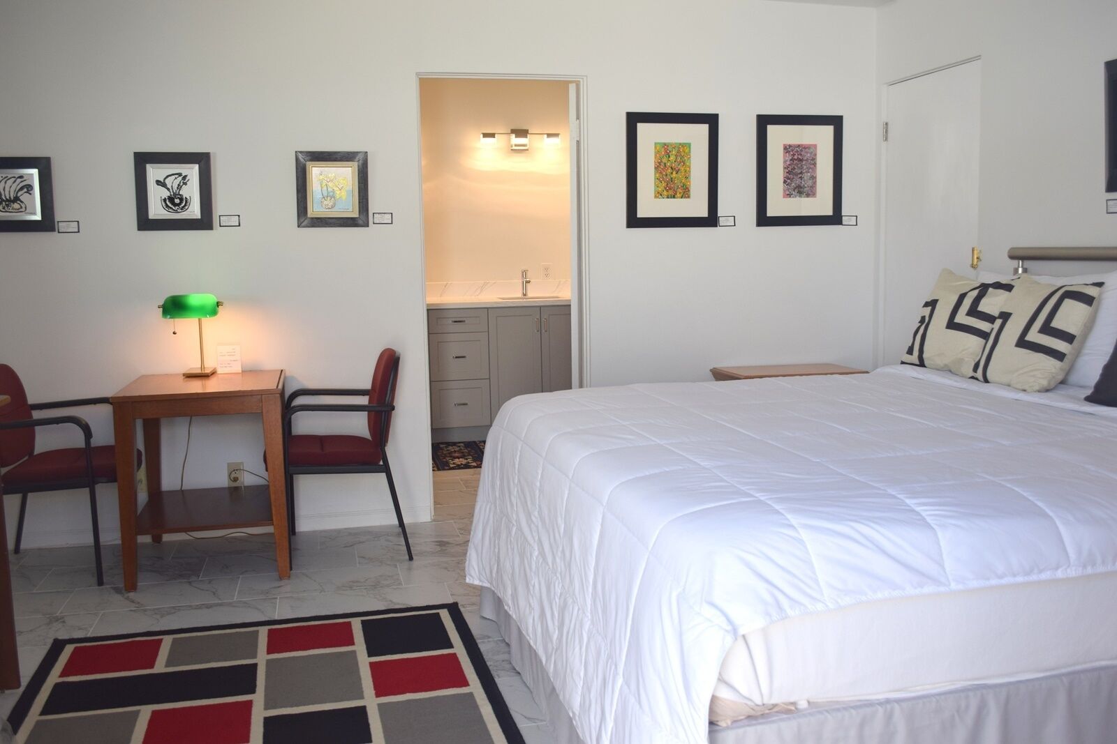 A bedroom with a bed, a desk with a chair, a small table with a lamp, and artwork on the walls.