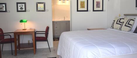 A bedroom with a bed, a desk with a chair, a small table with a lamp, and artwork on the walls.