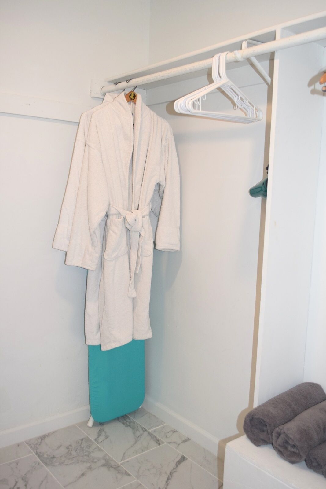 Une salle de bain avec un peignoir blanc suspendu à un porte-serviettes, un tapis de bain turquoise et une serviette grise posée sur un banc.