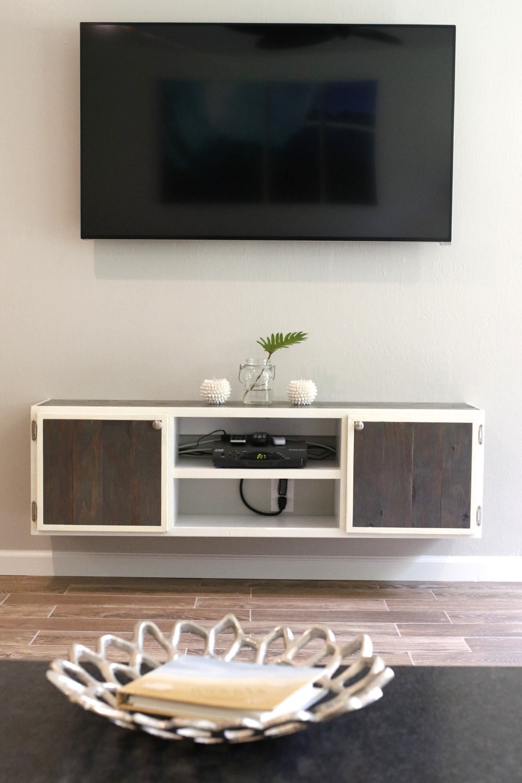 Un salon moderne avec un téléviseur à écran plat fixé au mur, un meuble en bois en dessous et un plateau décoratif posé sur le sol.