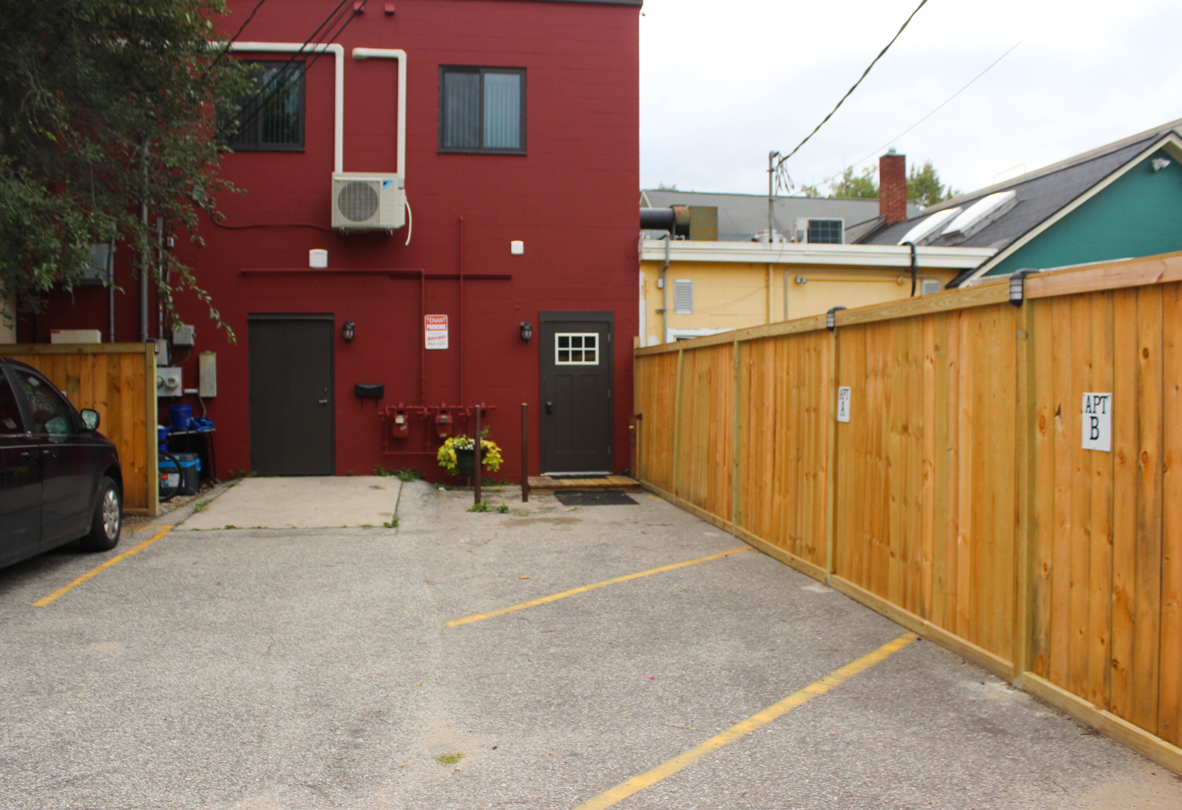 A parking area with a red building, a wooden fence, and a car parked on the left.