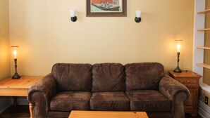 Uma sala de estar com sofá de couro marrom, mesa de centro de madeira, duas luminárias fixadas na parede e um quadro emoldurado.