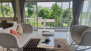 Una sala de estar moderna con sofá, puf y sillones, una mesa de centro y ventanas amplias con vistas a la vegetación.