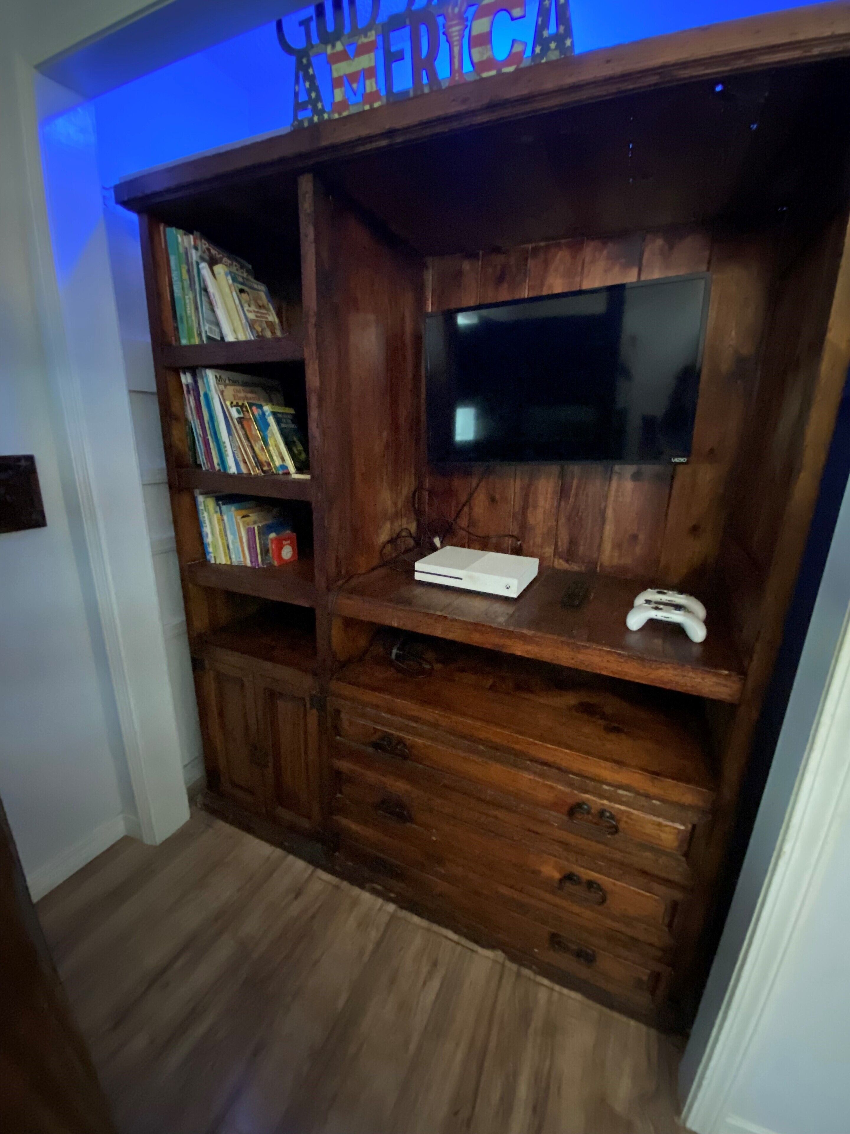 A wooden entertainment center with a flat-screen TV, a gaming console, and a bookshelf.