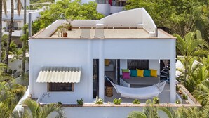 A modern two-story house with a flat roof, a hammock, and a balcony with colorful cushions.